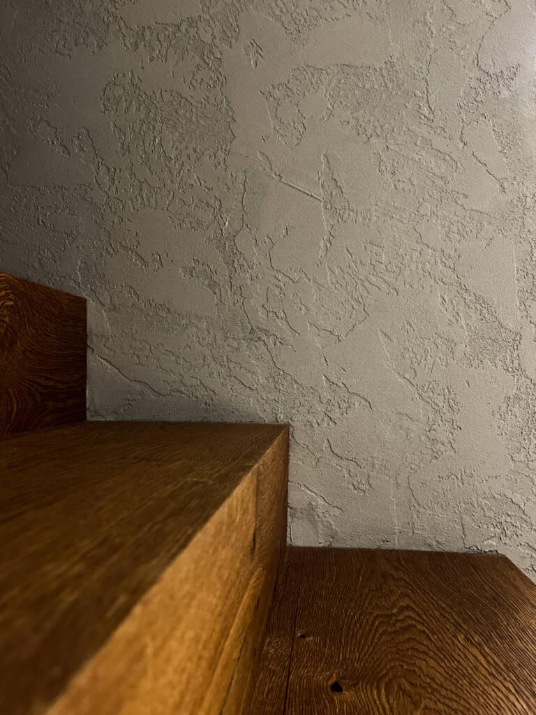 Parete con intonaco in argilla naturale a finitura materica accanto a scala in legno. Soluzione ecologica per bioedilizia.