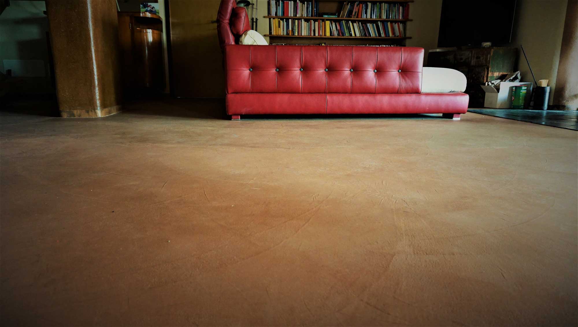 ClayPutty: il pavimento naturale che unisce estetica, tecnologia e sostenibilità 2 Vista ravvicinata di un pavimento in terra cruda color ocra con superficie liscia e texture naturale, sullo sfondo un divano rosso e una libreria.