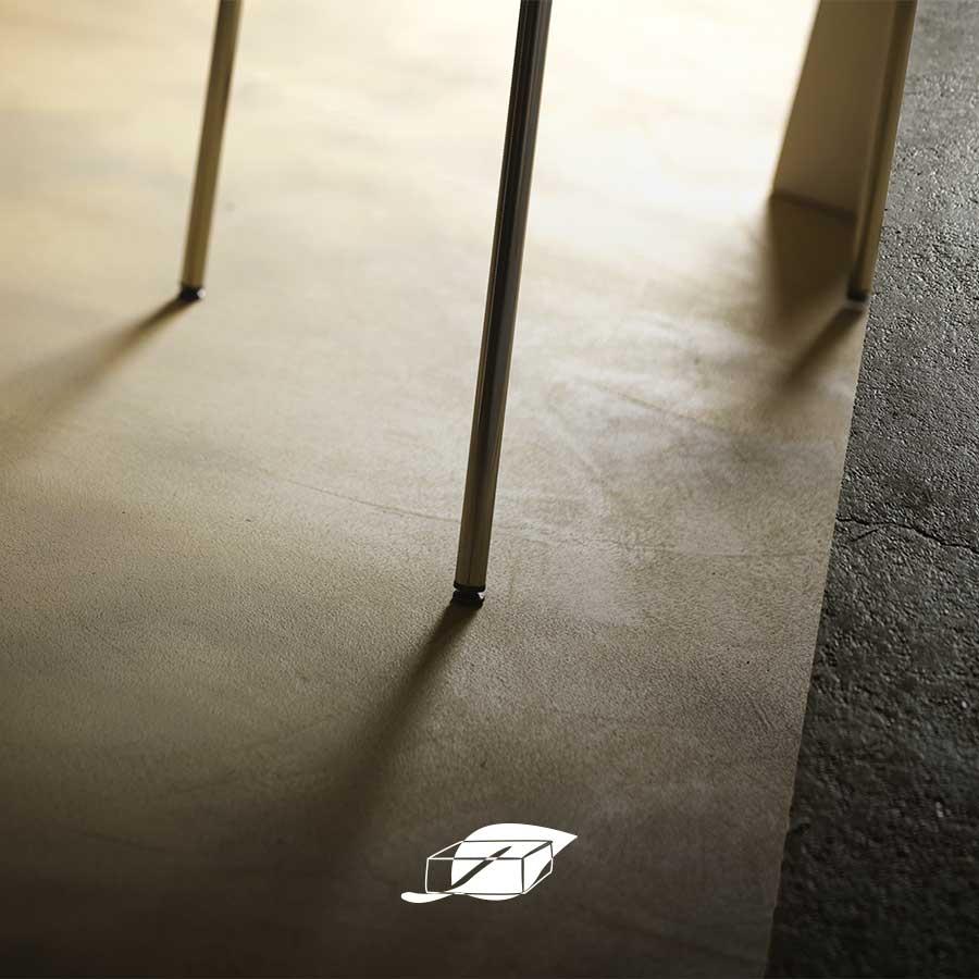 Dettaglio di un pavimento in terra cruda chiara accanto a una superficie in cemento grezzo, con ombre proiettate dalle gambe di una sedia.