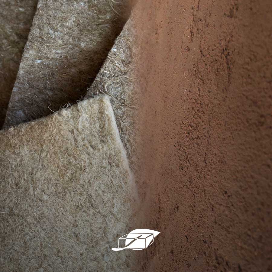 Rotolo di isolante in canapa appoggiato a una parete rifinita in intonaco di terra cruda: combinazione naturale per il comfort abitativo.