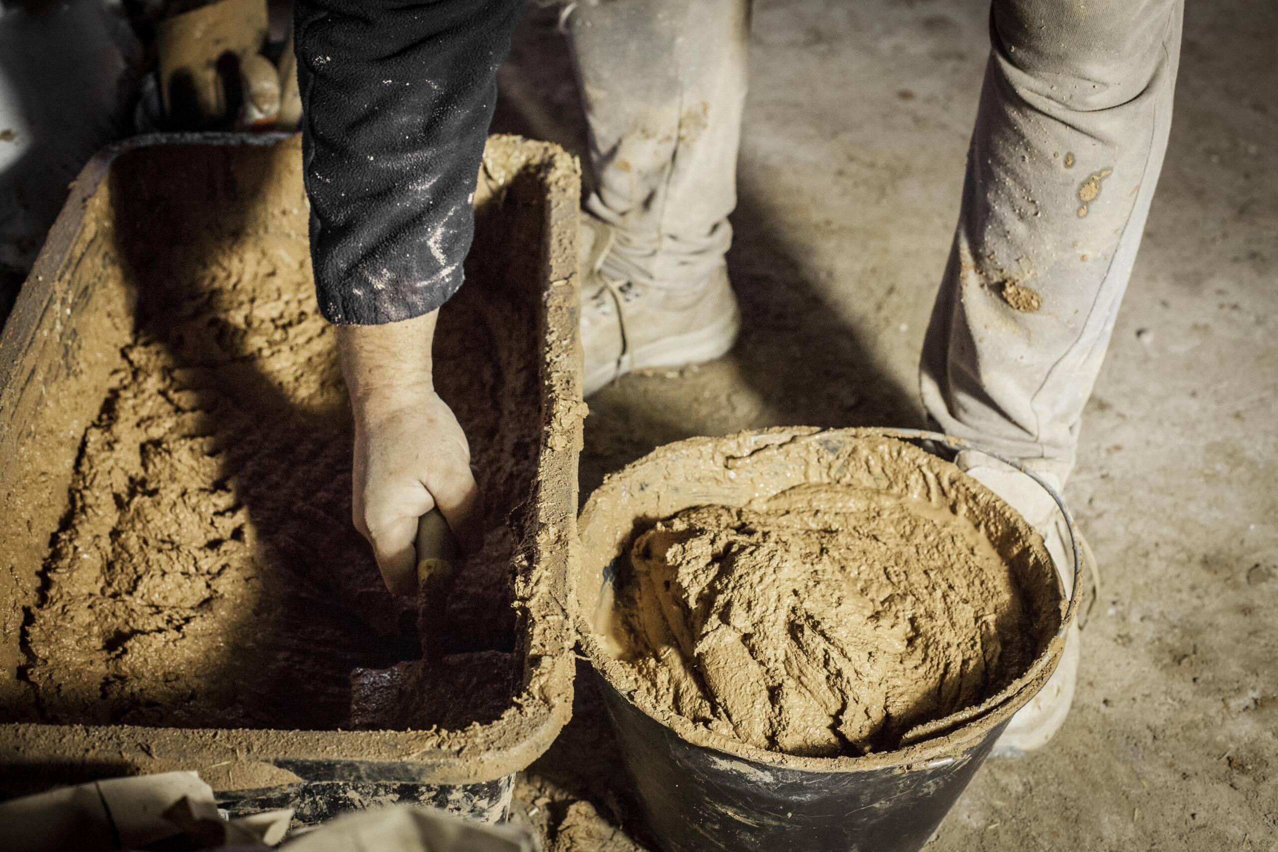 Due secchi pieni di argilla cruda pronta all’uso, con una mano che raccoglie il materiale con una spatola per iniziare la stesura su una superficie.