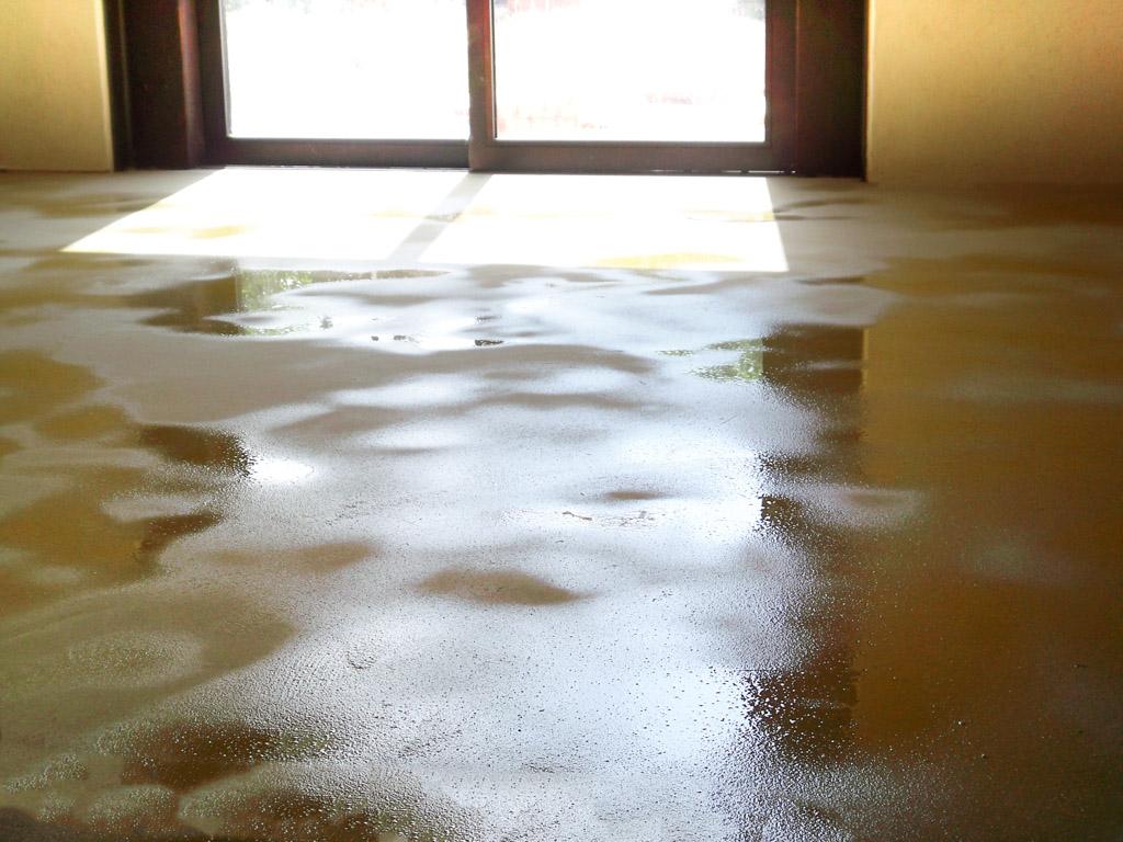 Pavimento in terra cruda appena steso, lucido e ancora umido, con riflessi di luce naturale vicino a una finestra.