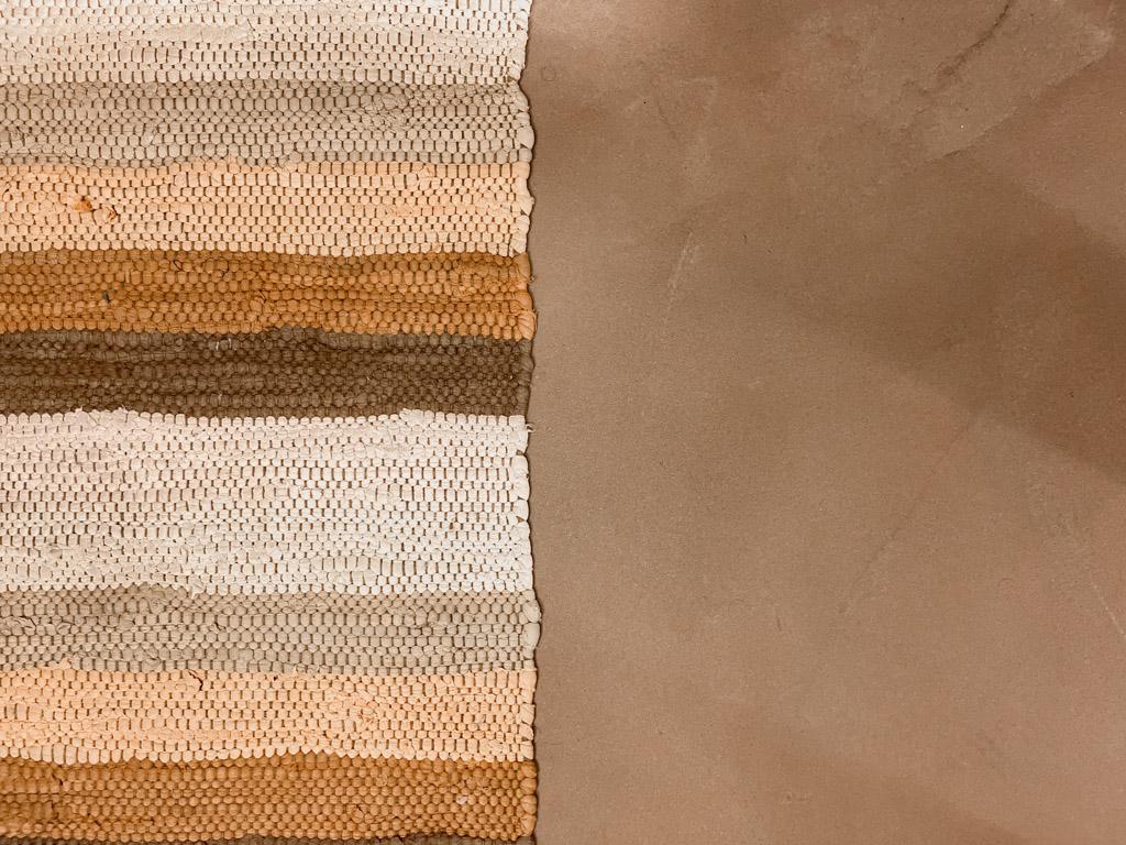 Superficie asciutta di un pavimento in argilla cruda accanto a un tappeto intrecciato multicolore, che mostra la texture materica.